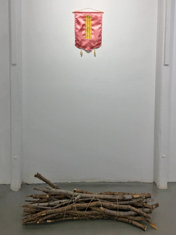 On the wall is an embroidered tapestry on a pink satin background of a bundle of sticks. On the ground in front of the wall is an actual bundle of sticks.