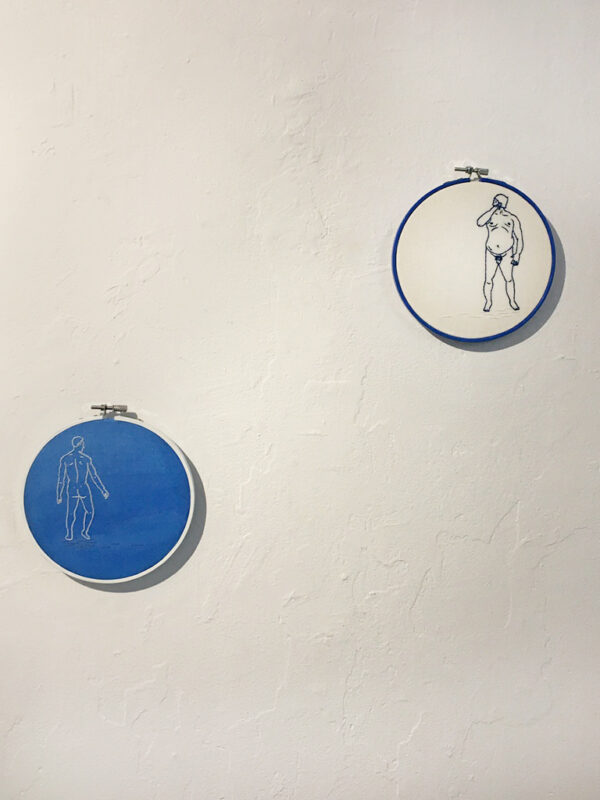 Two embroidered pieces on a wall, the bottom left one is of a male figure in a white line on a blue background and the top left one is of a mal figure in a blue line on a white background.