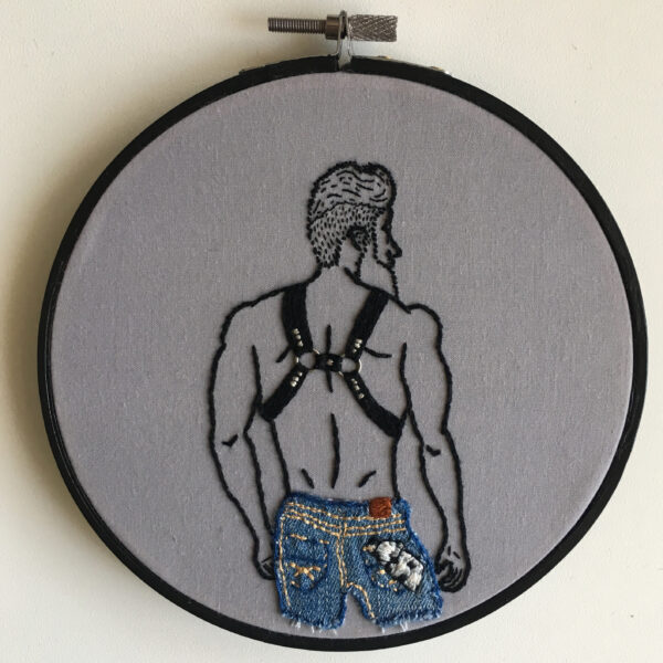 An embroidered piece of a the back of a man wearing a leather harness and jeans, with a black and white hanky sticking out of his right pocket.